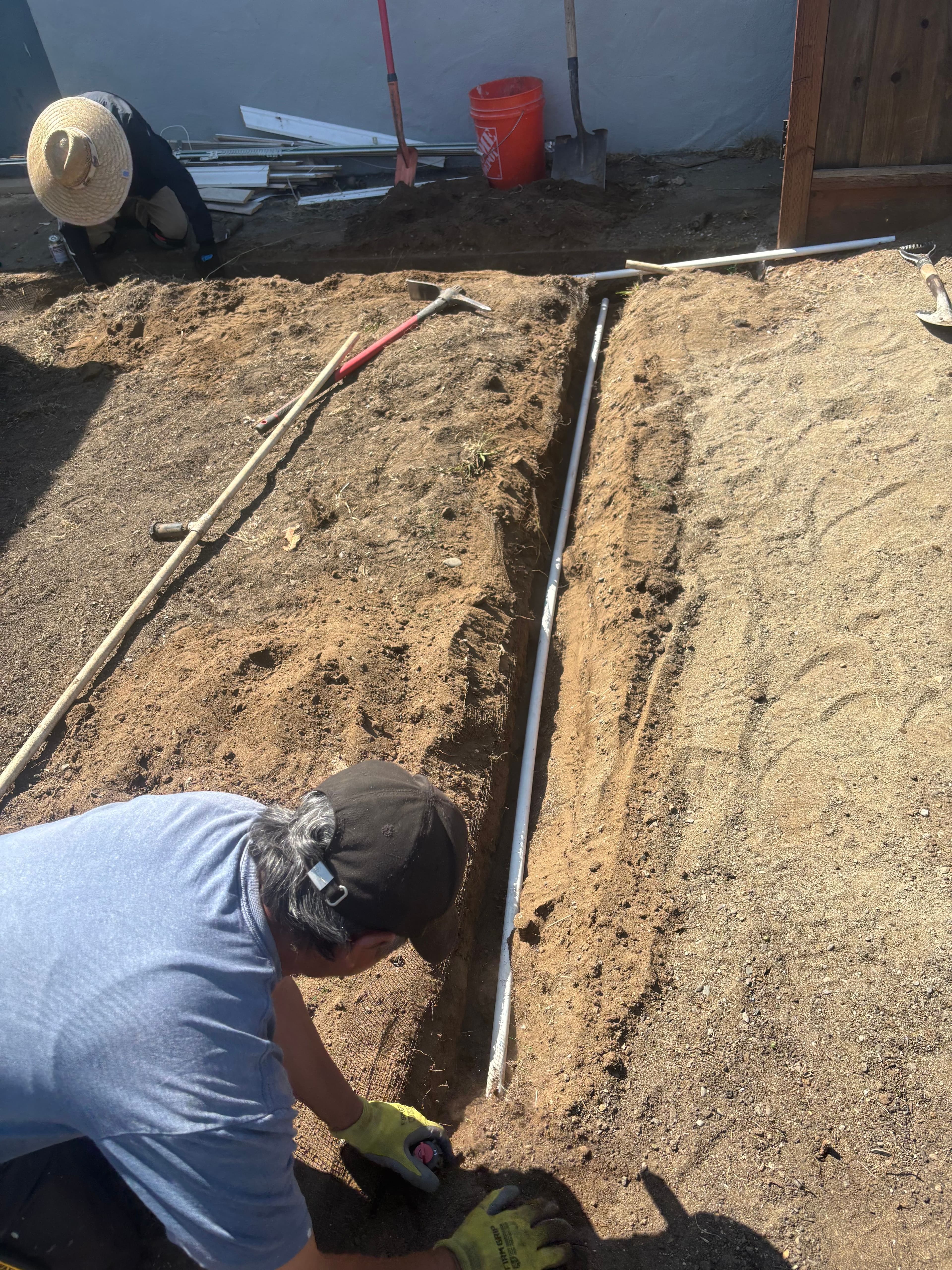 Two workers digging a trench in sandy soil for landscaping in a residential yard.