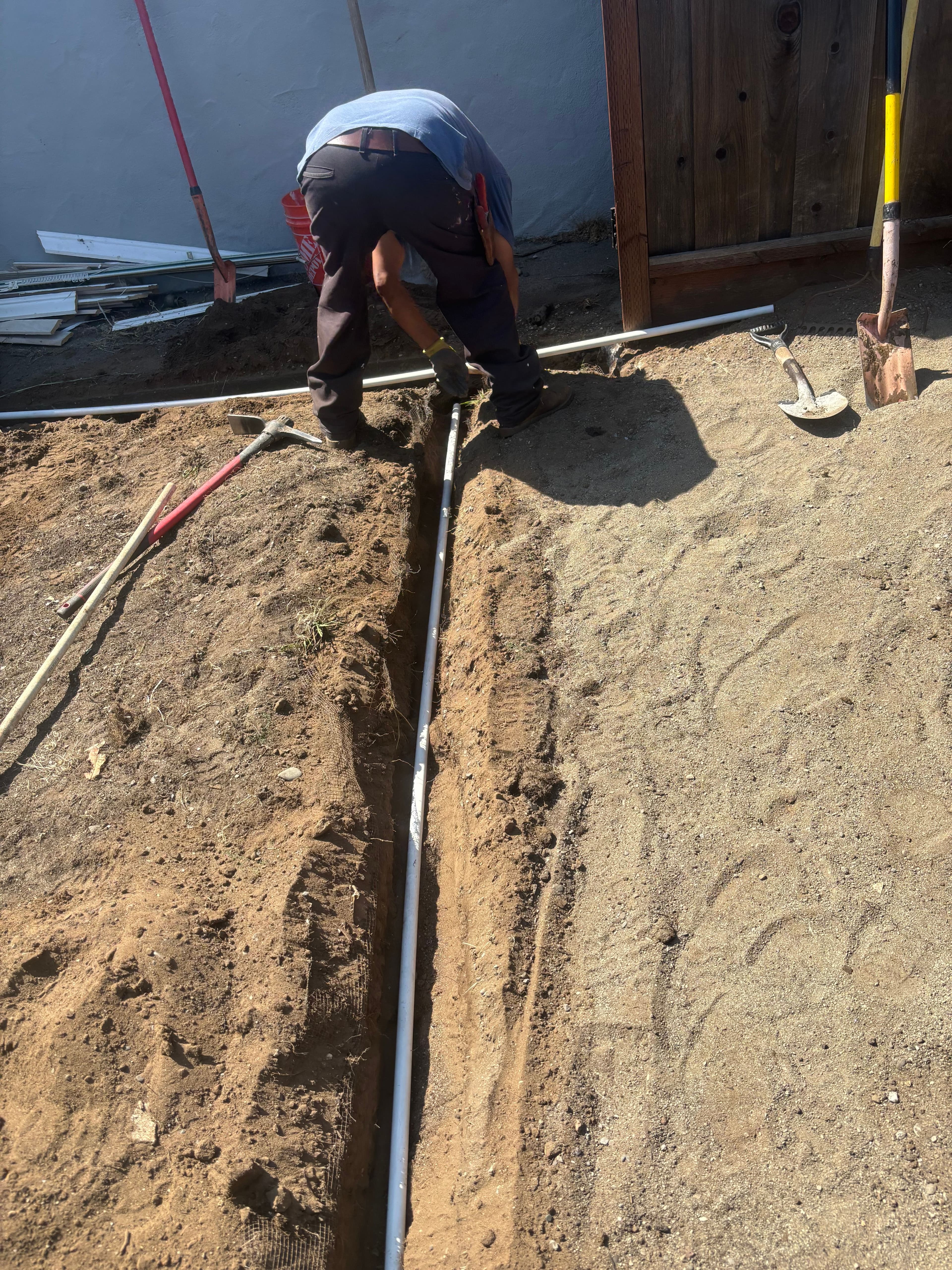 Worker installing irrigation pipe in a sandy trench outdoors with tools nearby.