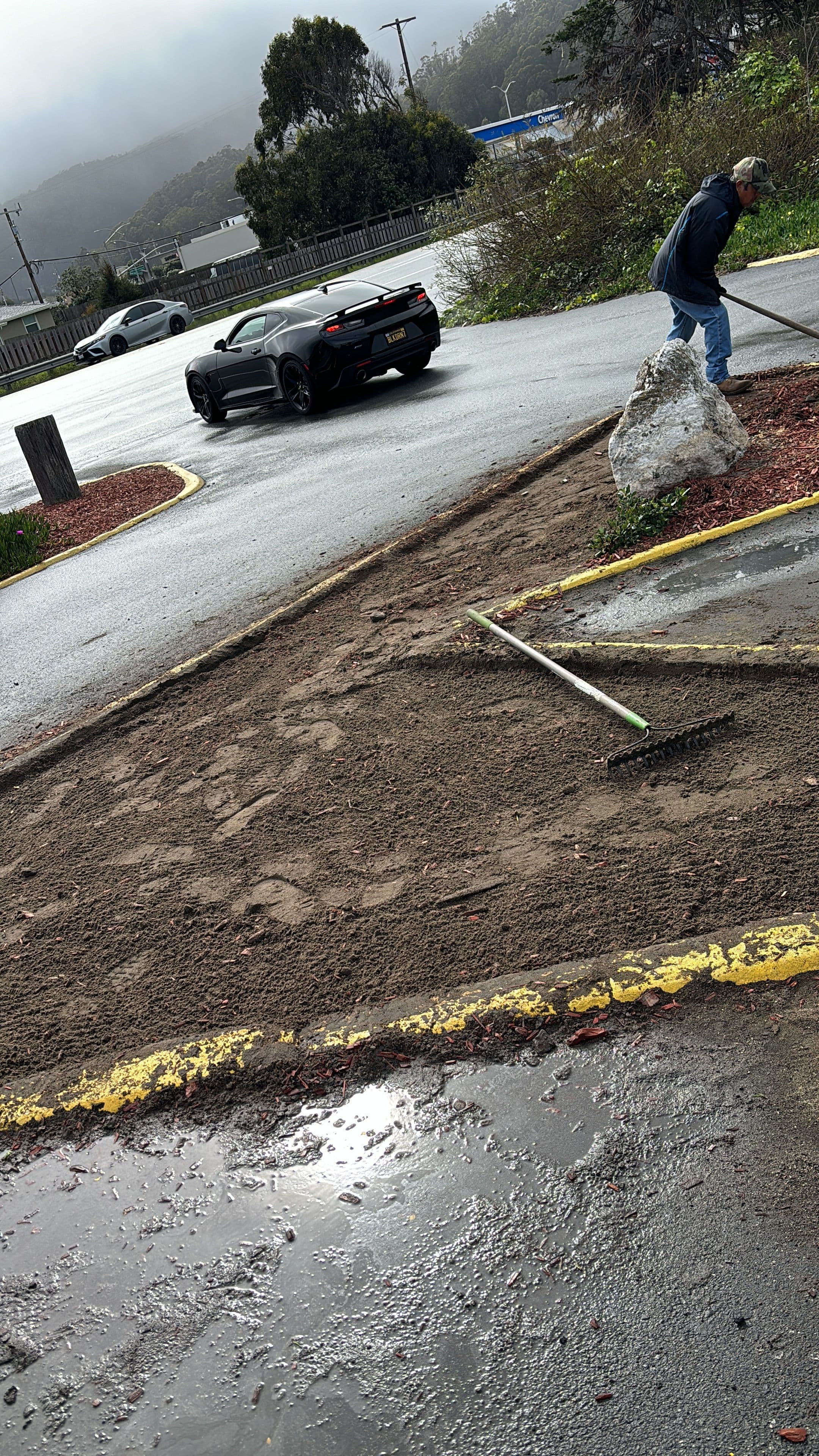 Man raking dirt in a parking lot with a car in the background on a rainy day.