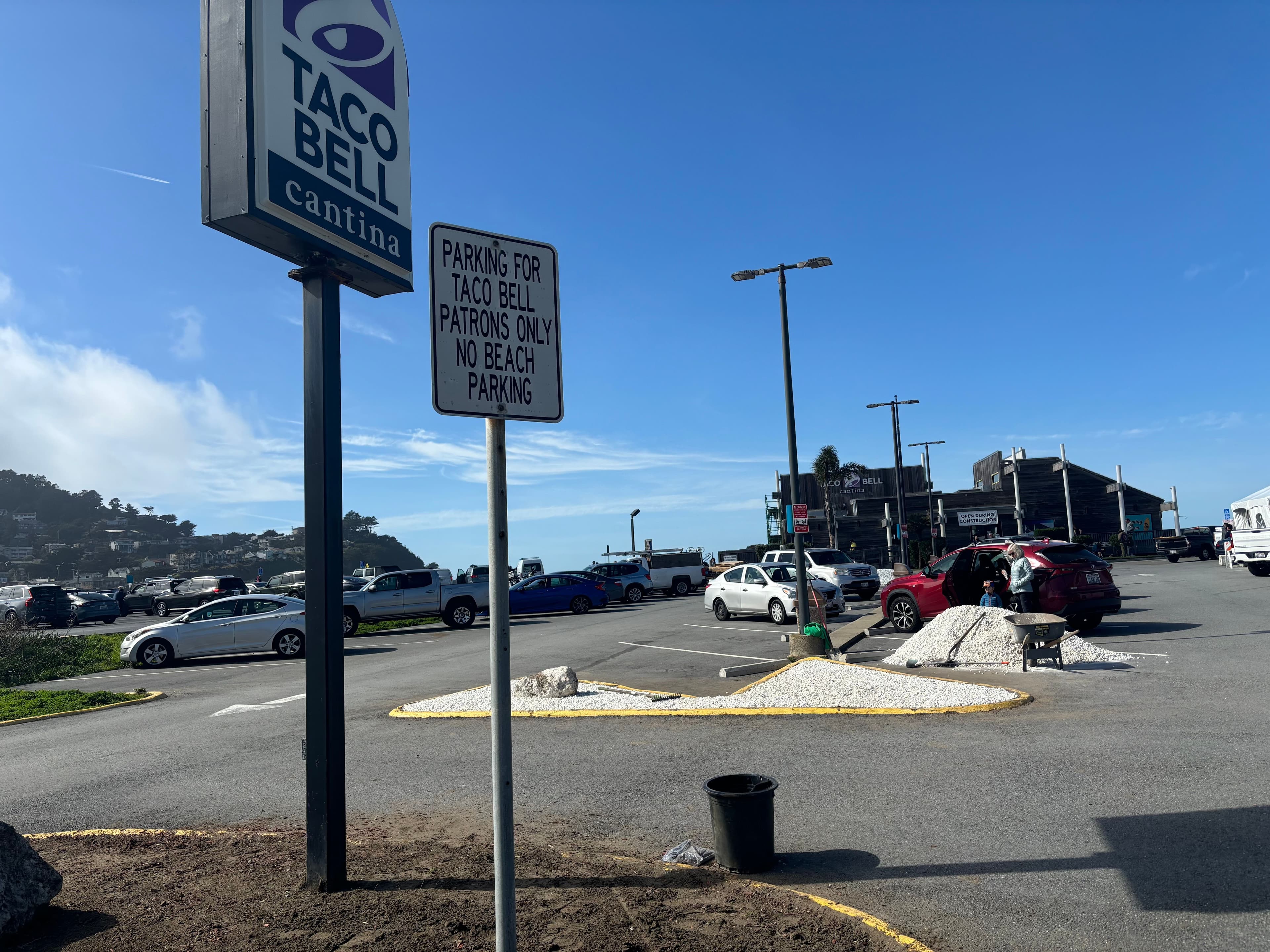 Taco Bell Cantina sign with parking lot, cars, and beach area in sunny coastal location.