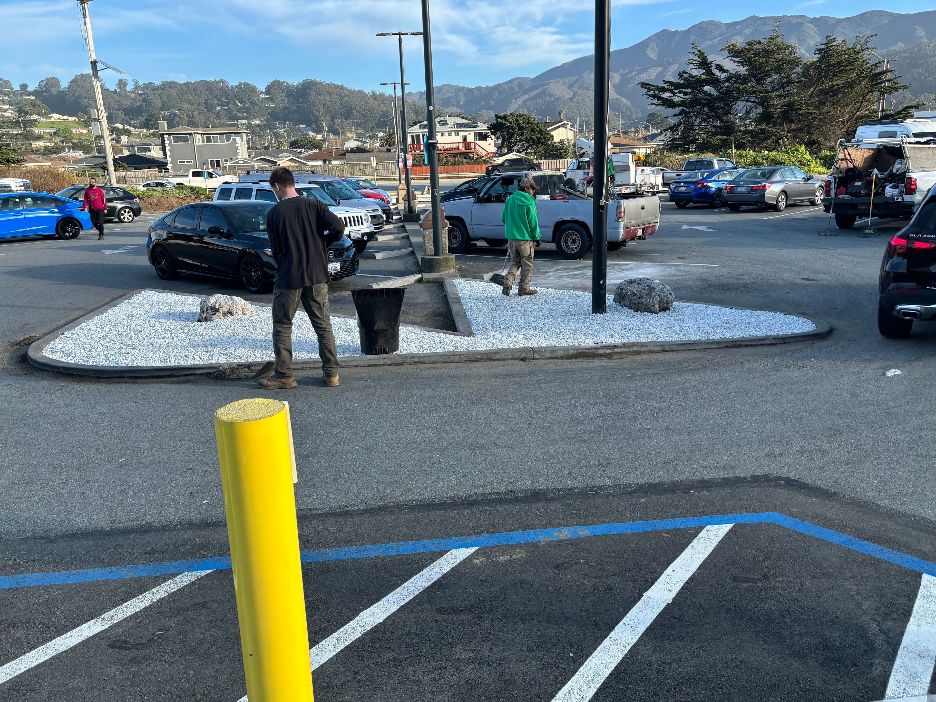 Parking lot scene with two men working near a landscaped area and parked cars in the background.