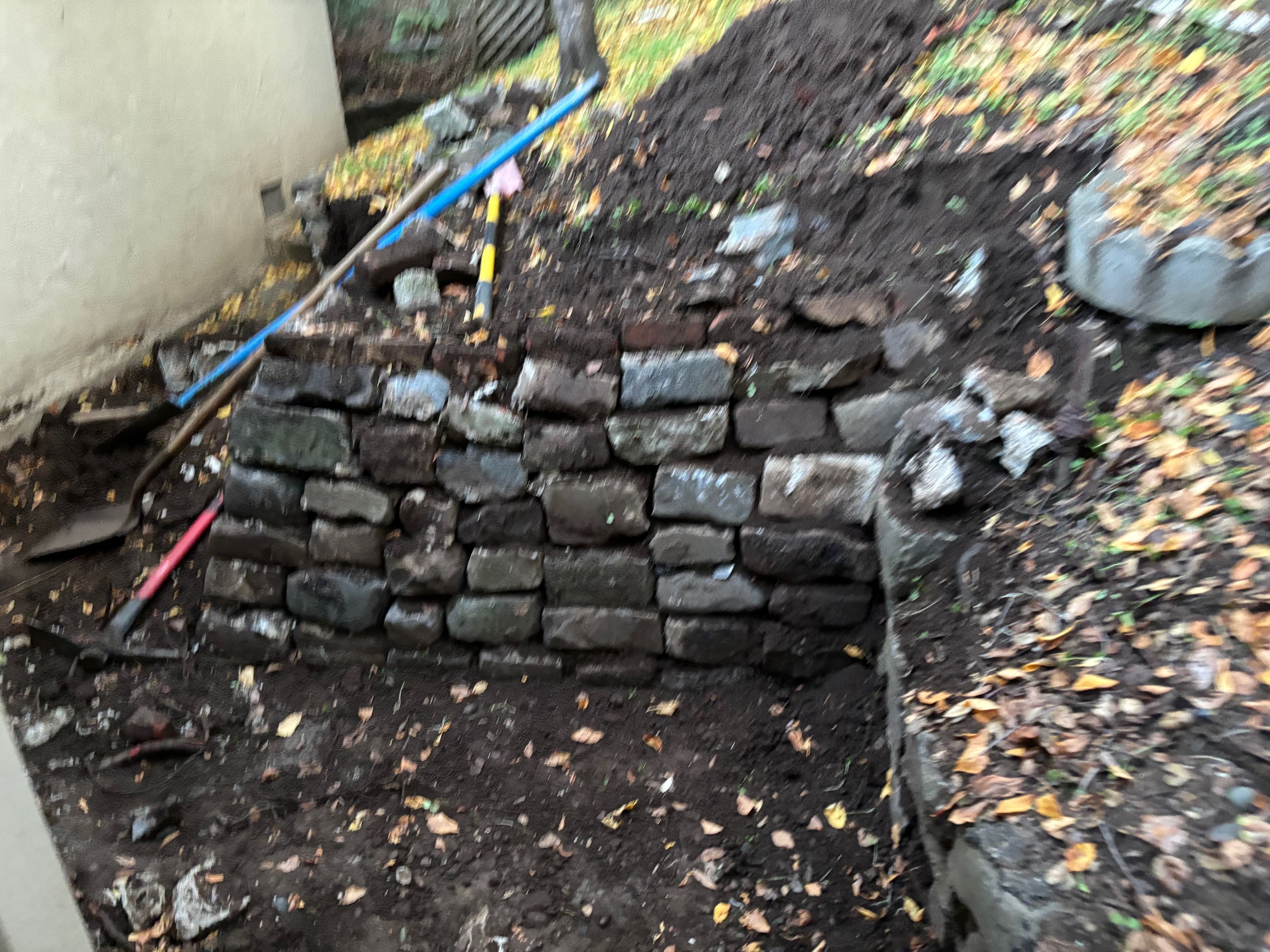 Stone wall construction in a garden, surrounded by dirt and fallen leaves. Tools visible.