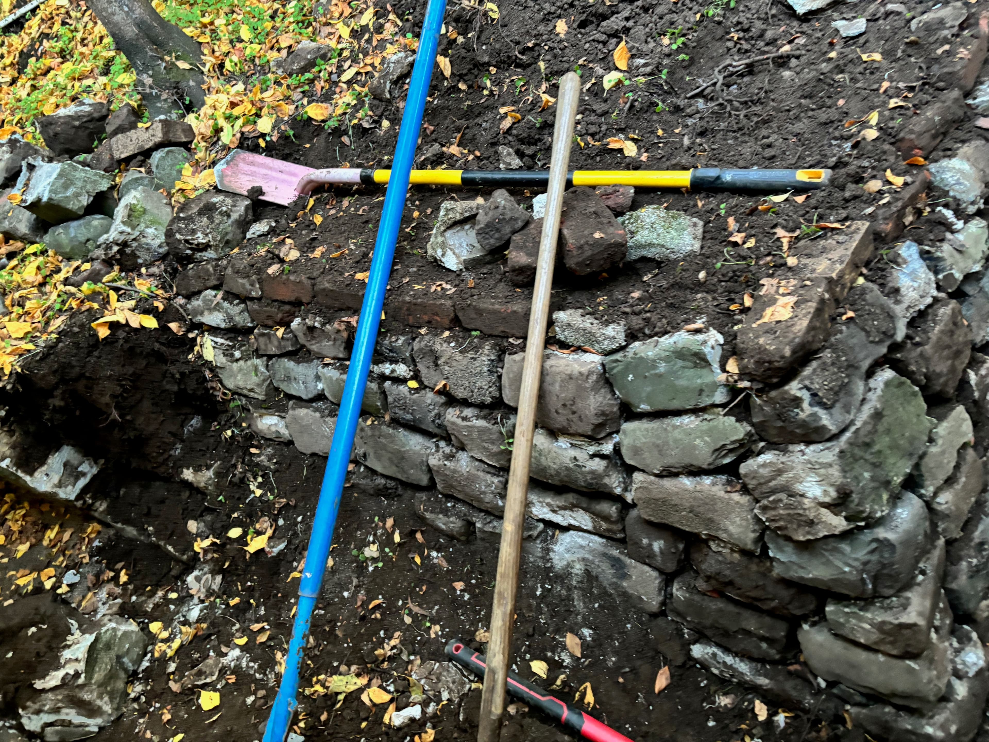 Stonewall construction project with tools like a shovel and a wooden post on earthy ground.