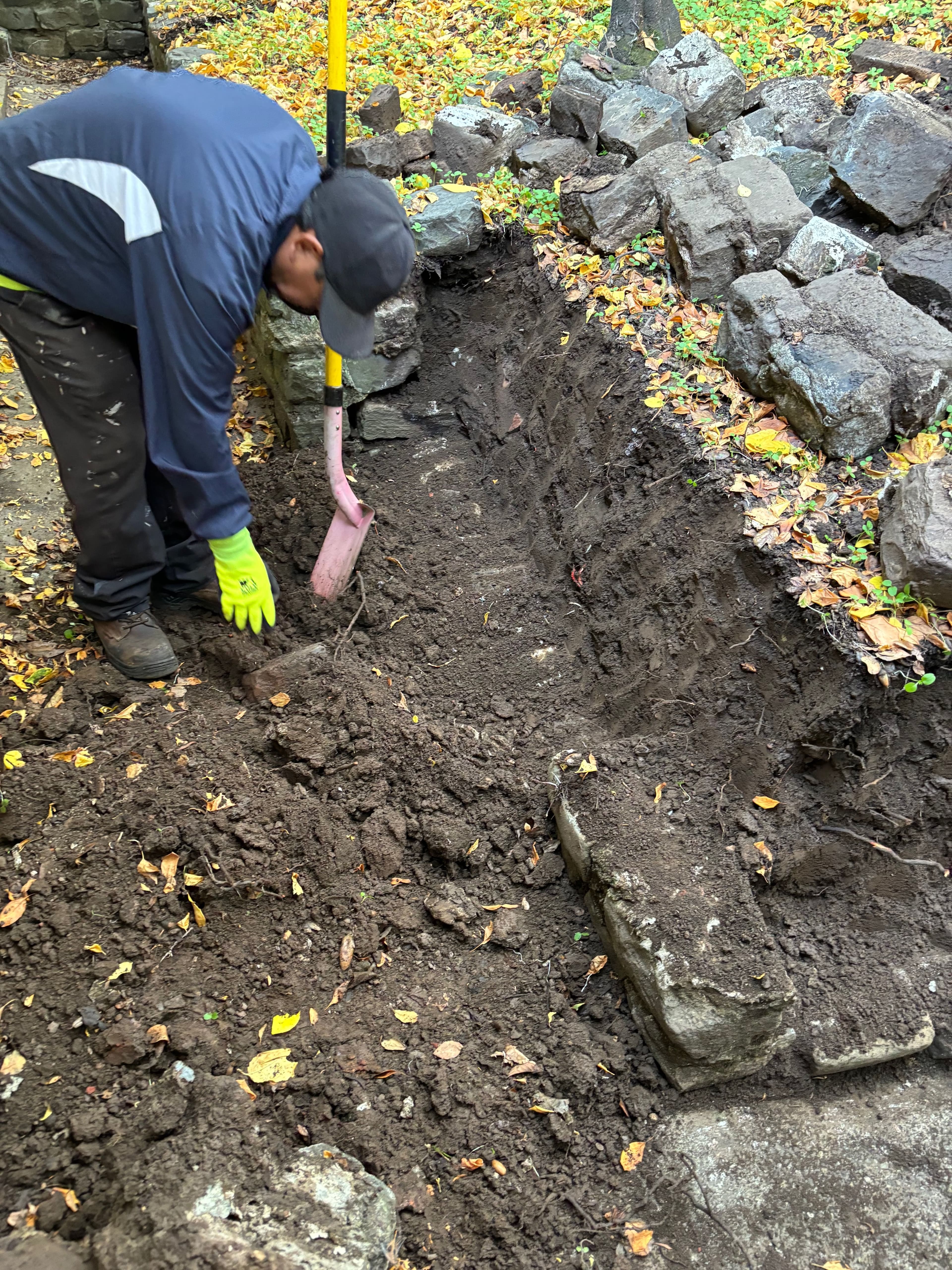 Person digging a trench in a wooded area, surrounded by rocks and autumn leaves.
