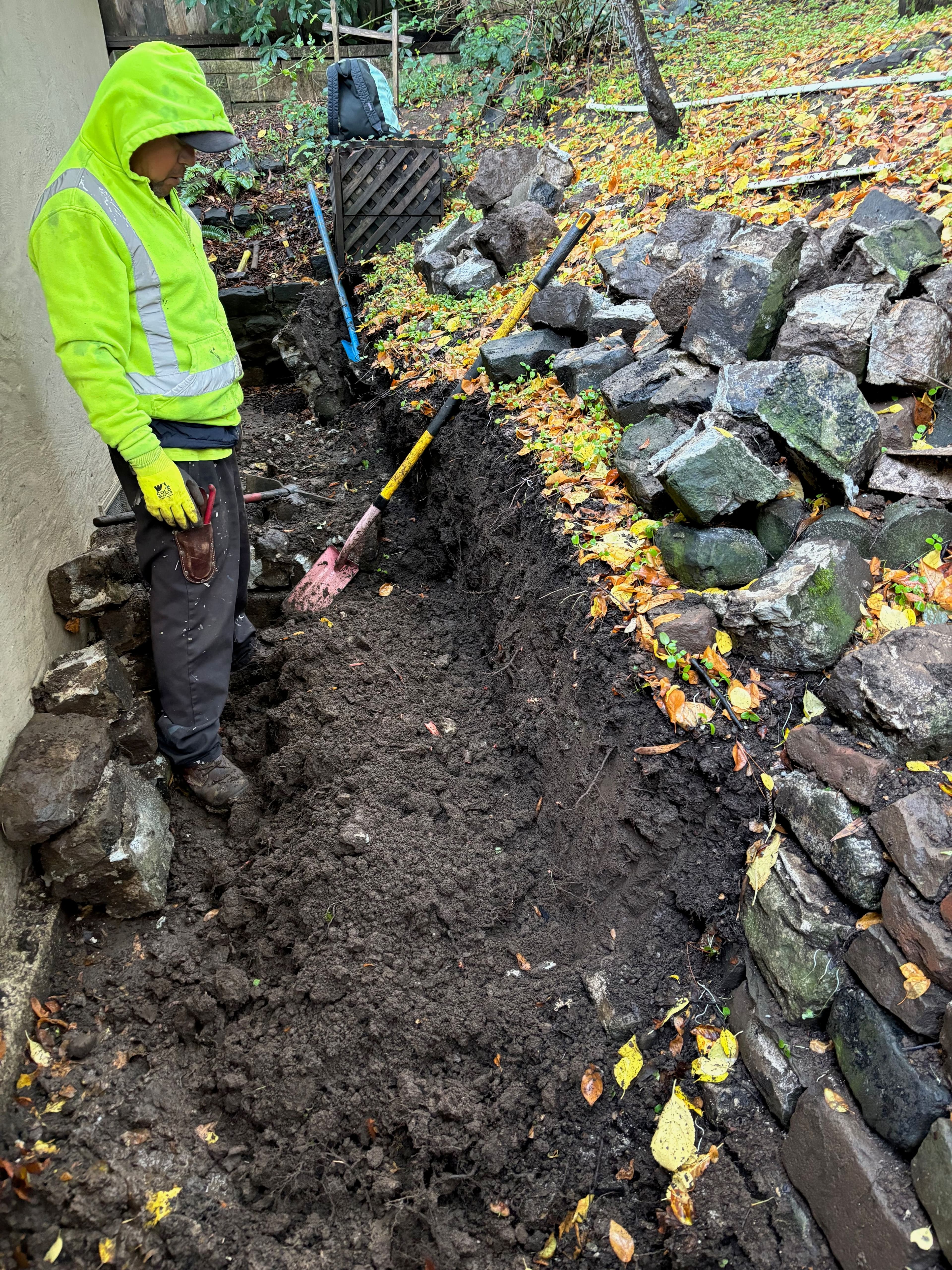 Construction worker digging a trench in a garden with stone walls and autumn leaves.