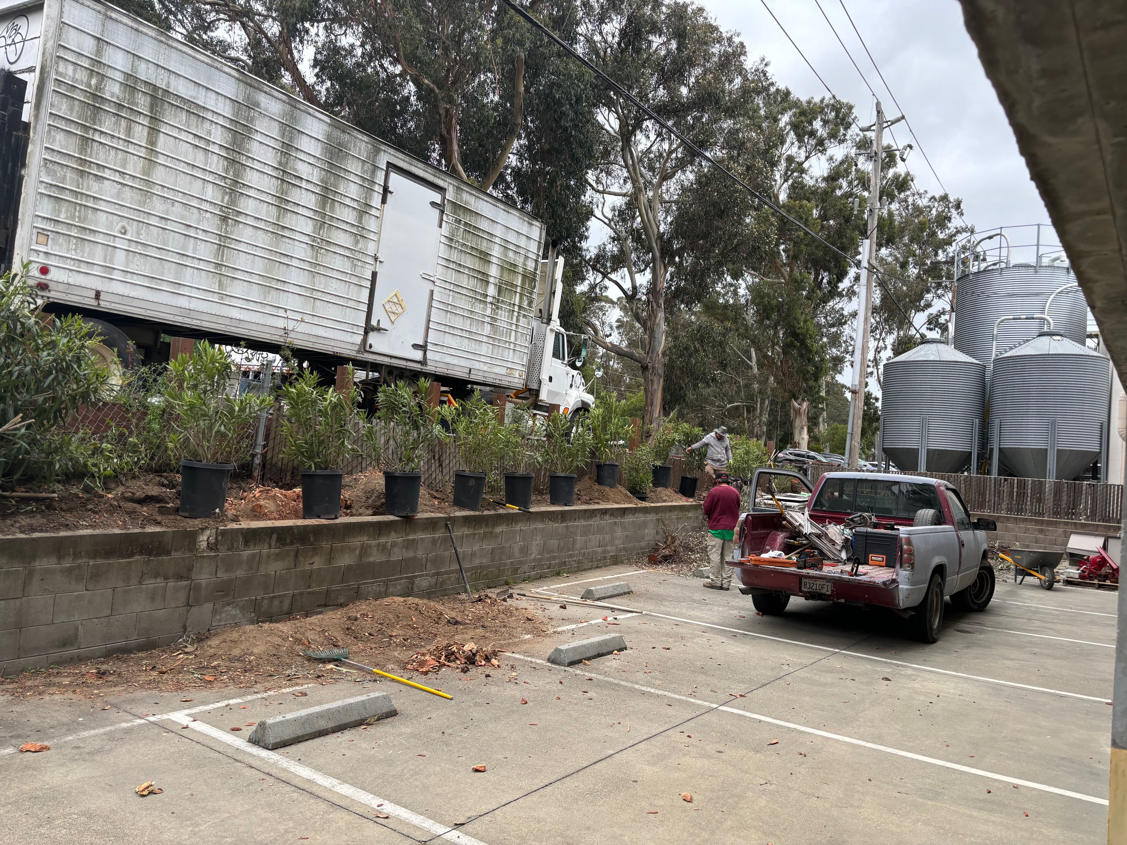 Truck unloading near a parking lot with a red pickup and planter boxes in a commercial area.