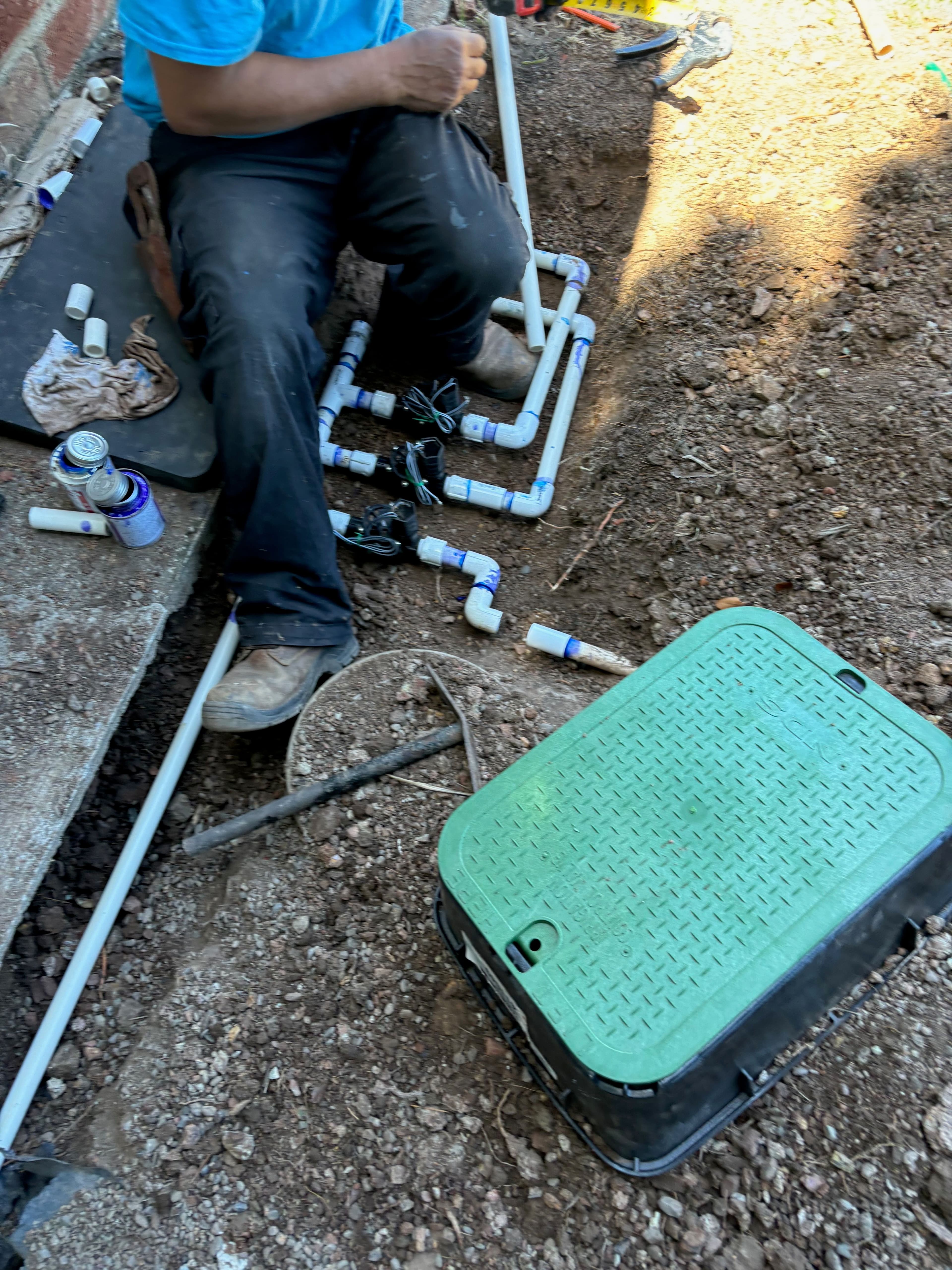 Person working on PVC pipe installation in a dug-out area with tools and materials nearby.