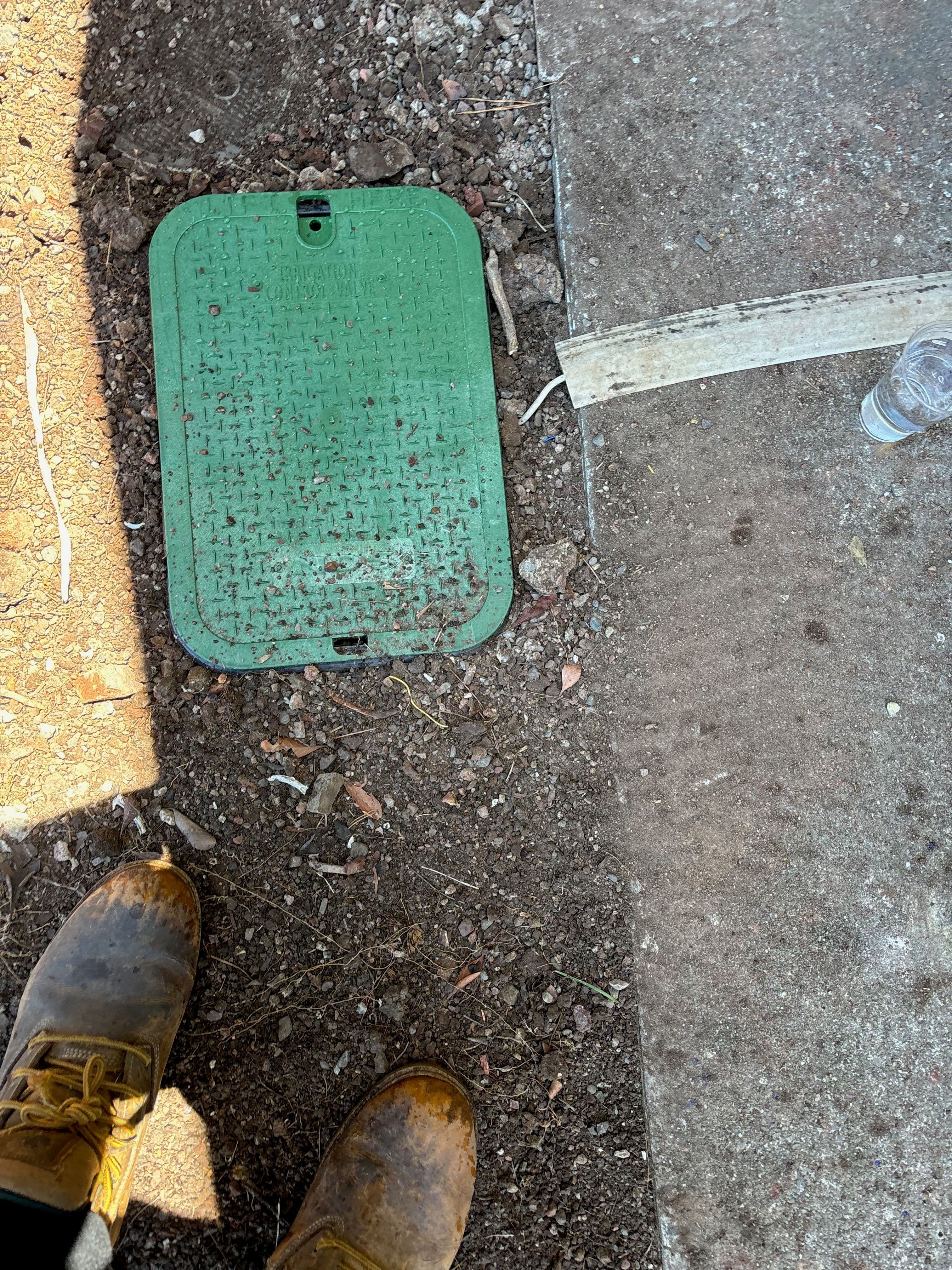 Green utility cover on dirt ground with work boots nearby.