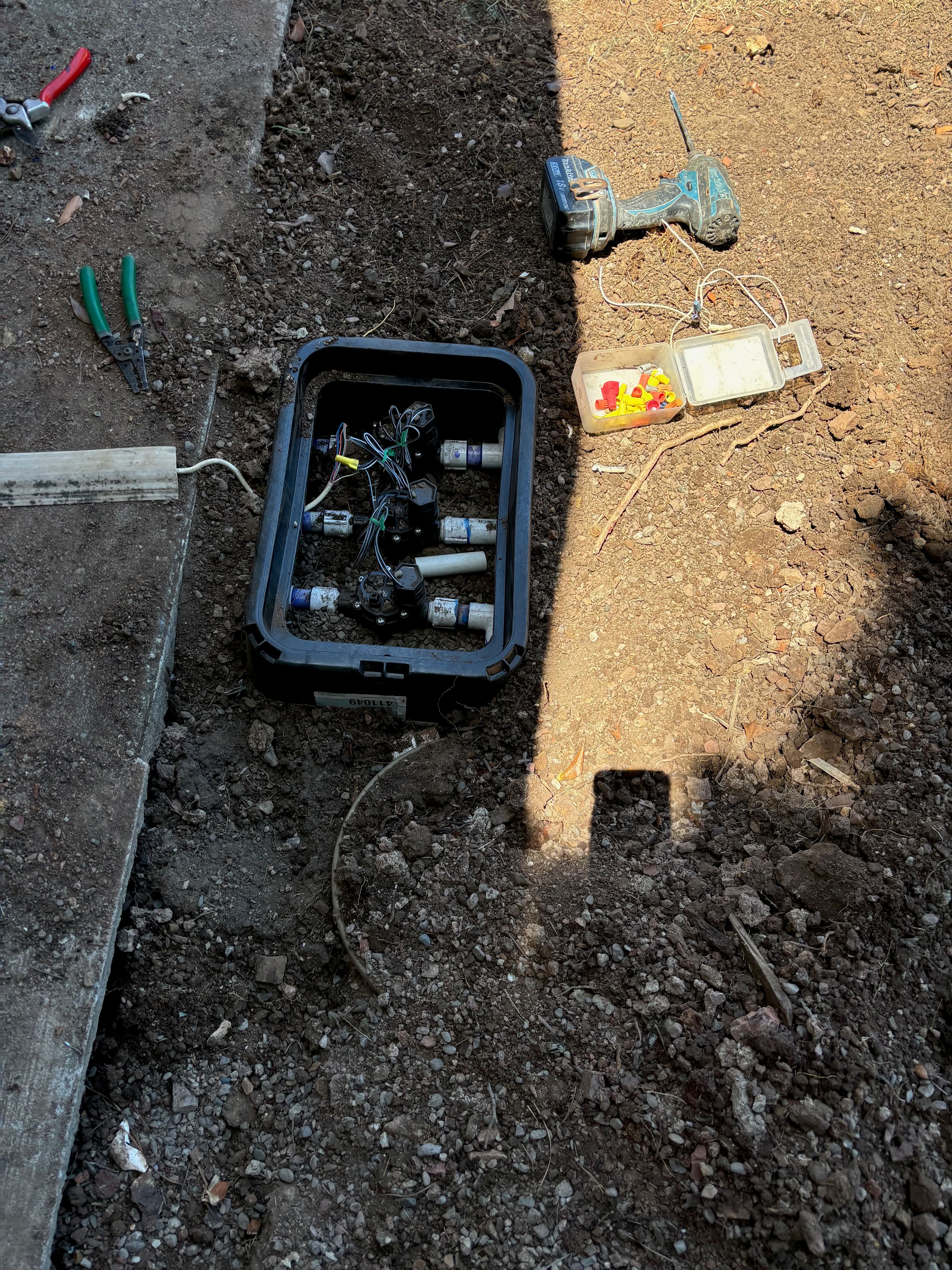 Wiring tools and a junction box with connections on a gravel surface in sunlight.