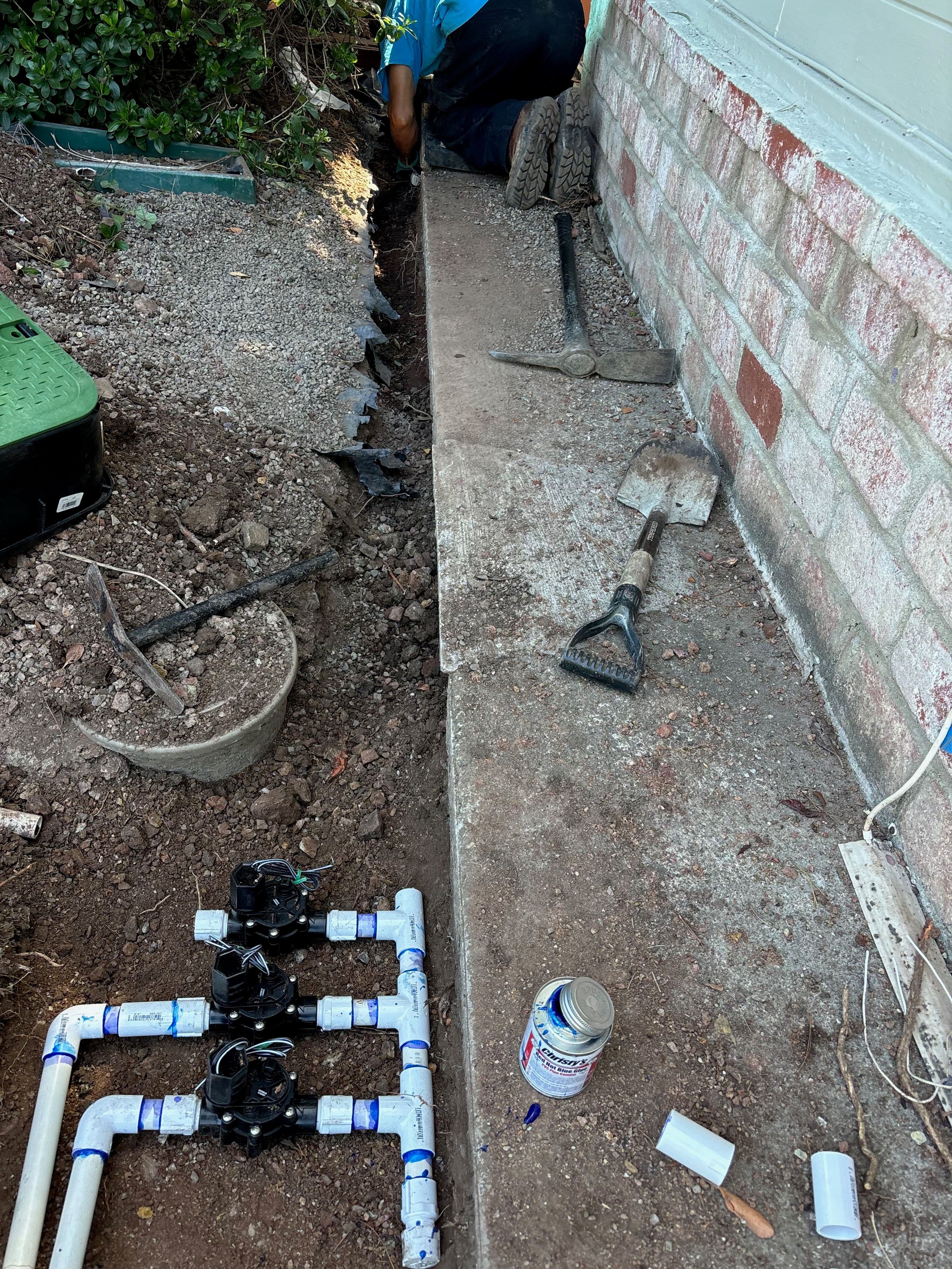 Worker installing irrigation system with PVC pipes and tools near a home's foundation.