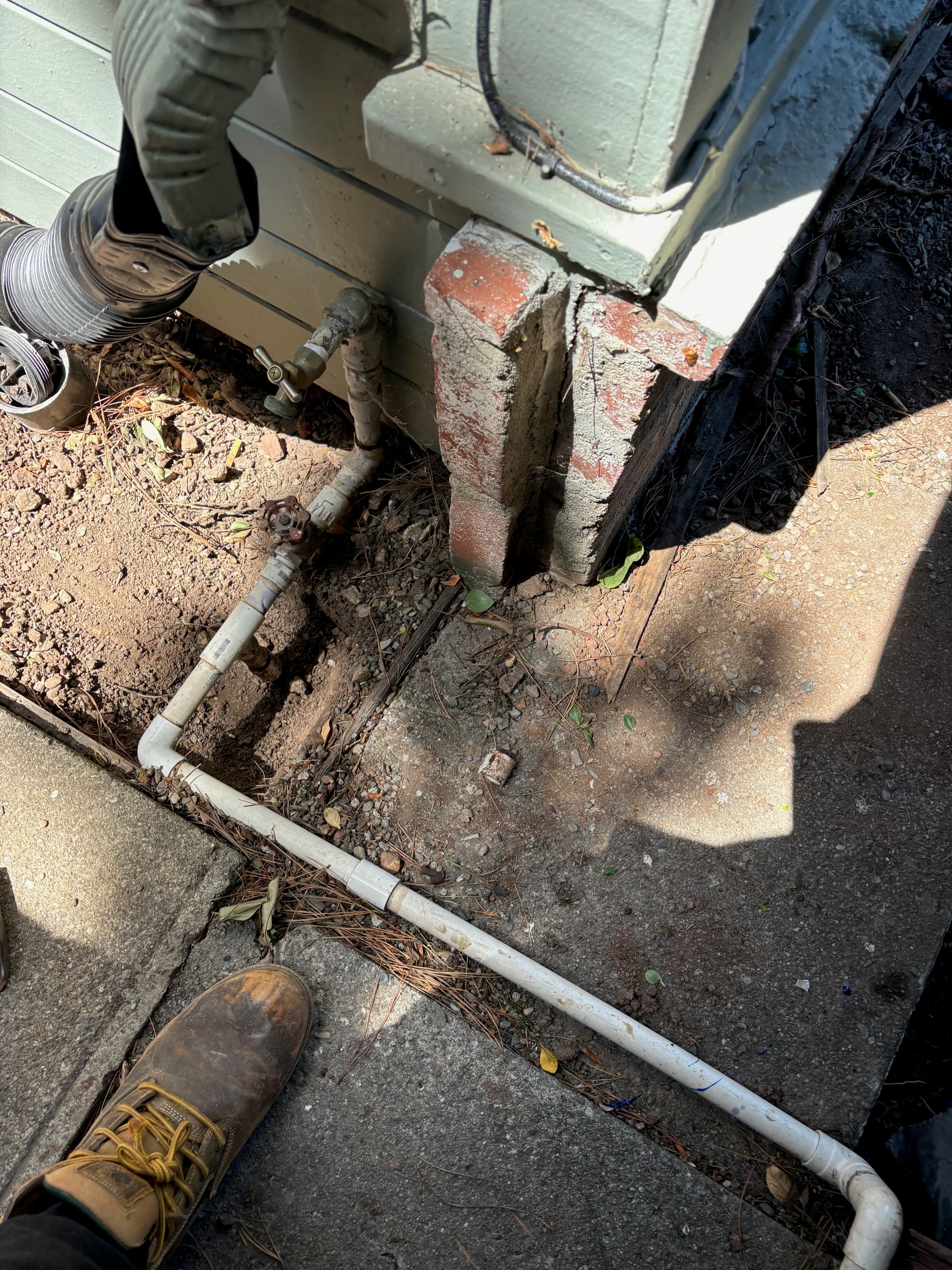 Plumbing pipes and connections near a building foundation with a worker's boot in view.