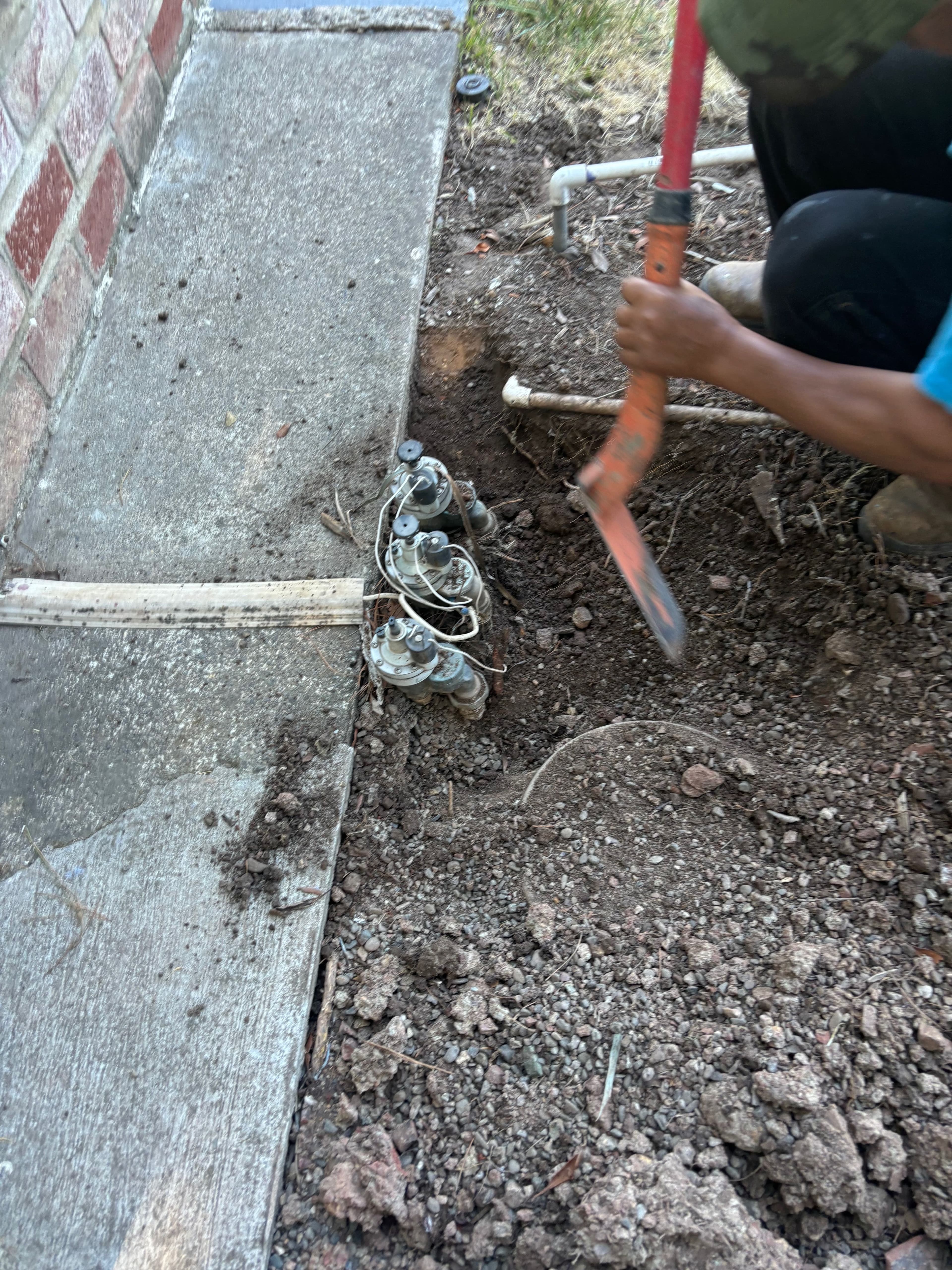 Person digging near a brick wall, exposing plumbing components in the soil.