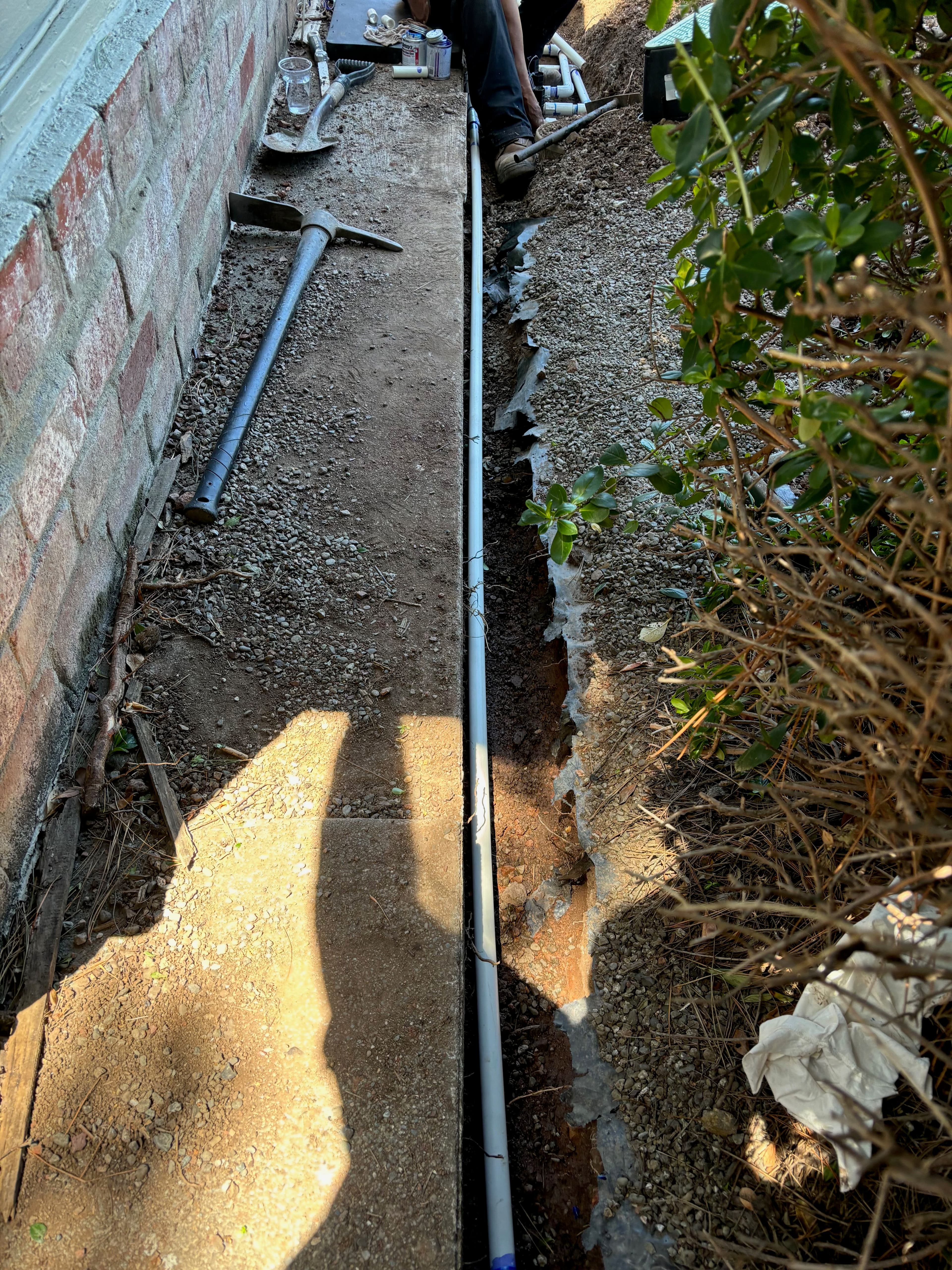 Trench for plumbing installation beside a brick wall, with tools and plants nearby.