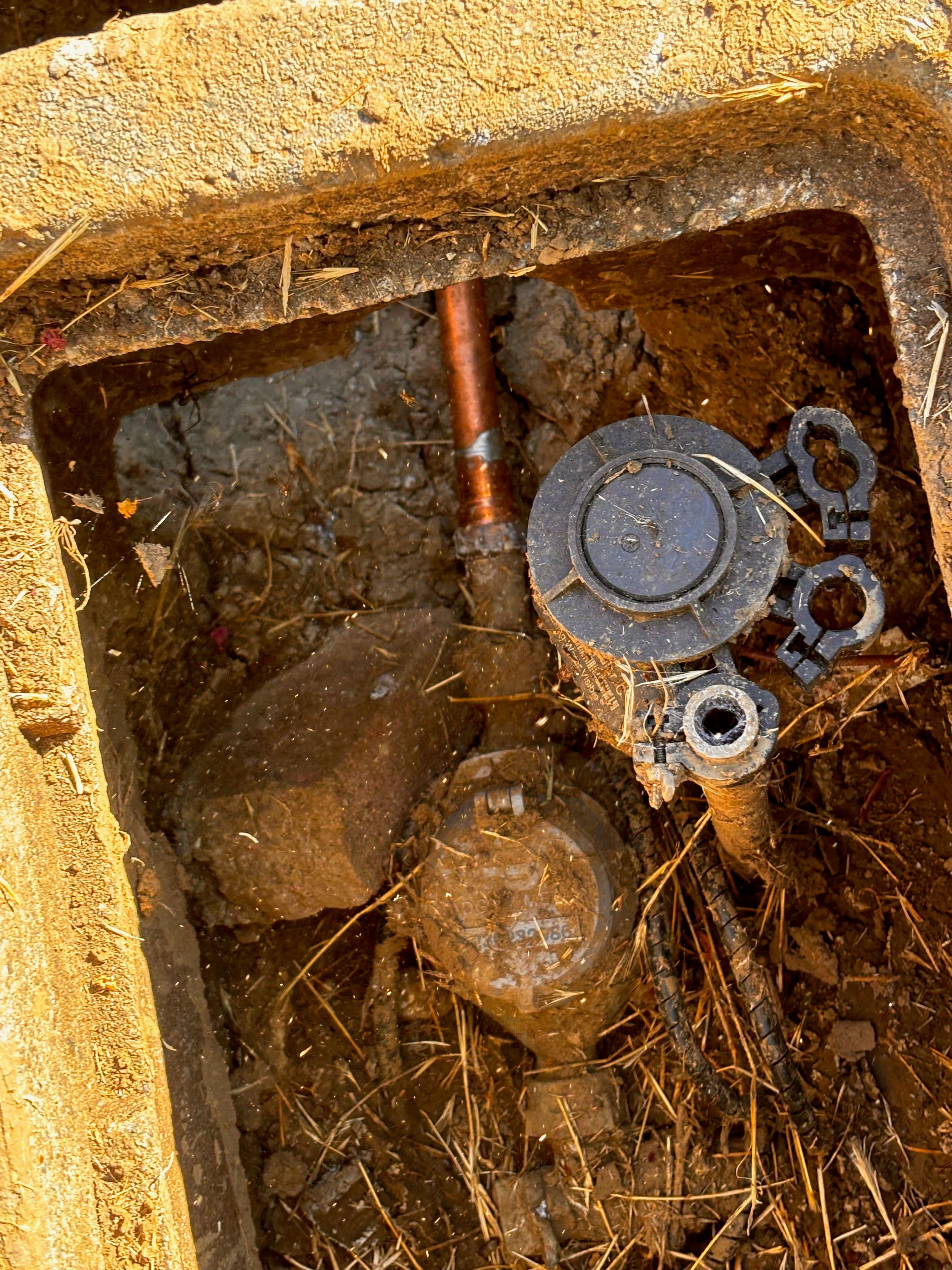Water meter and valve in a dirt-covered access pit, surrounded by debris and soil.