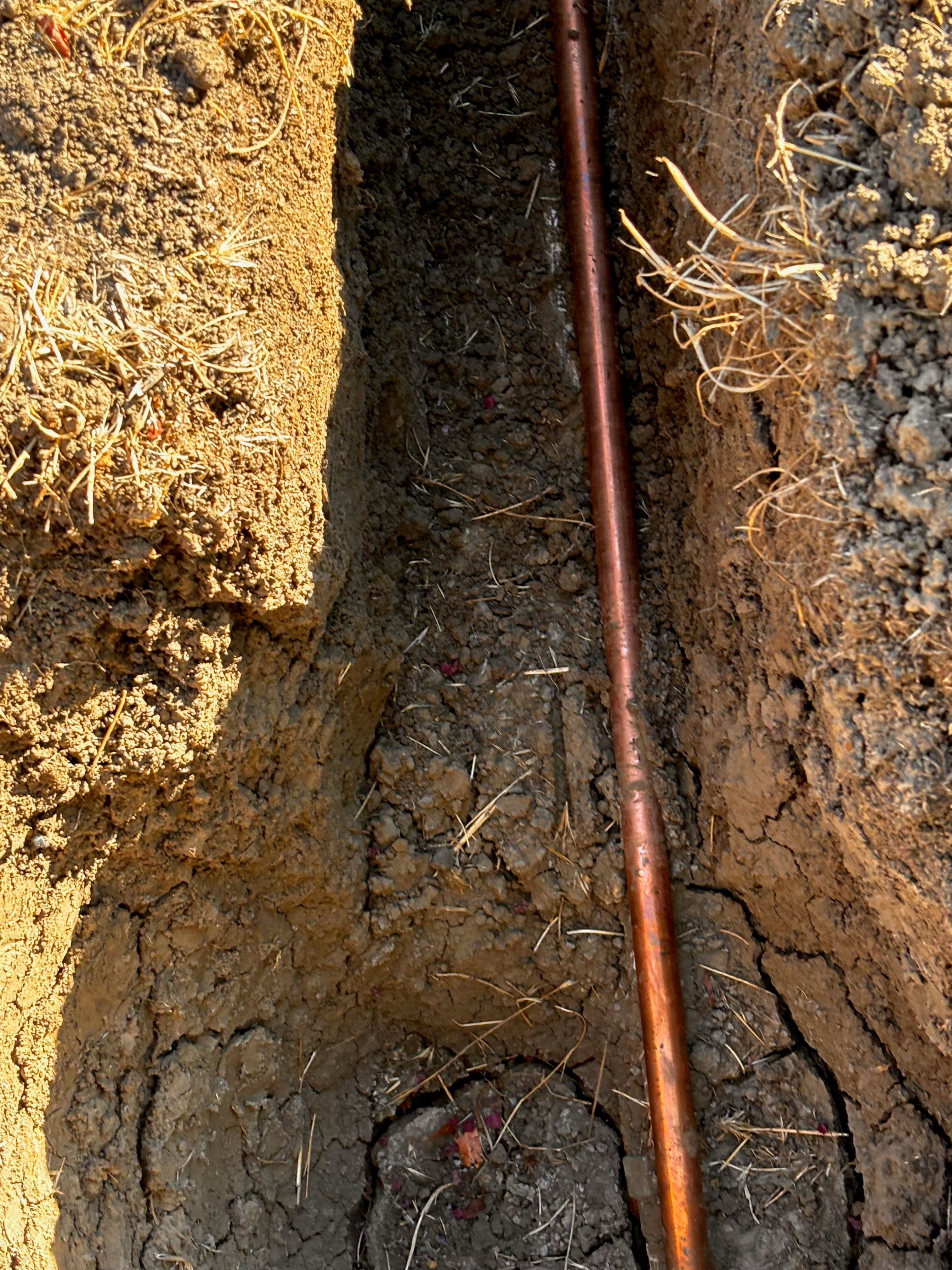 Copper pipe laid in an excavation trench surrounded by loose earth and dry grass.