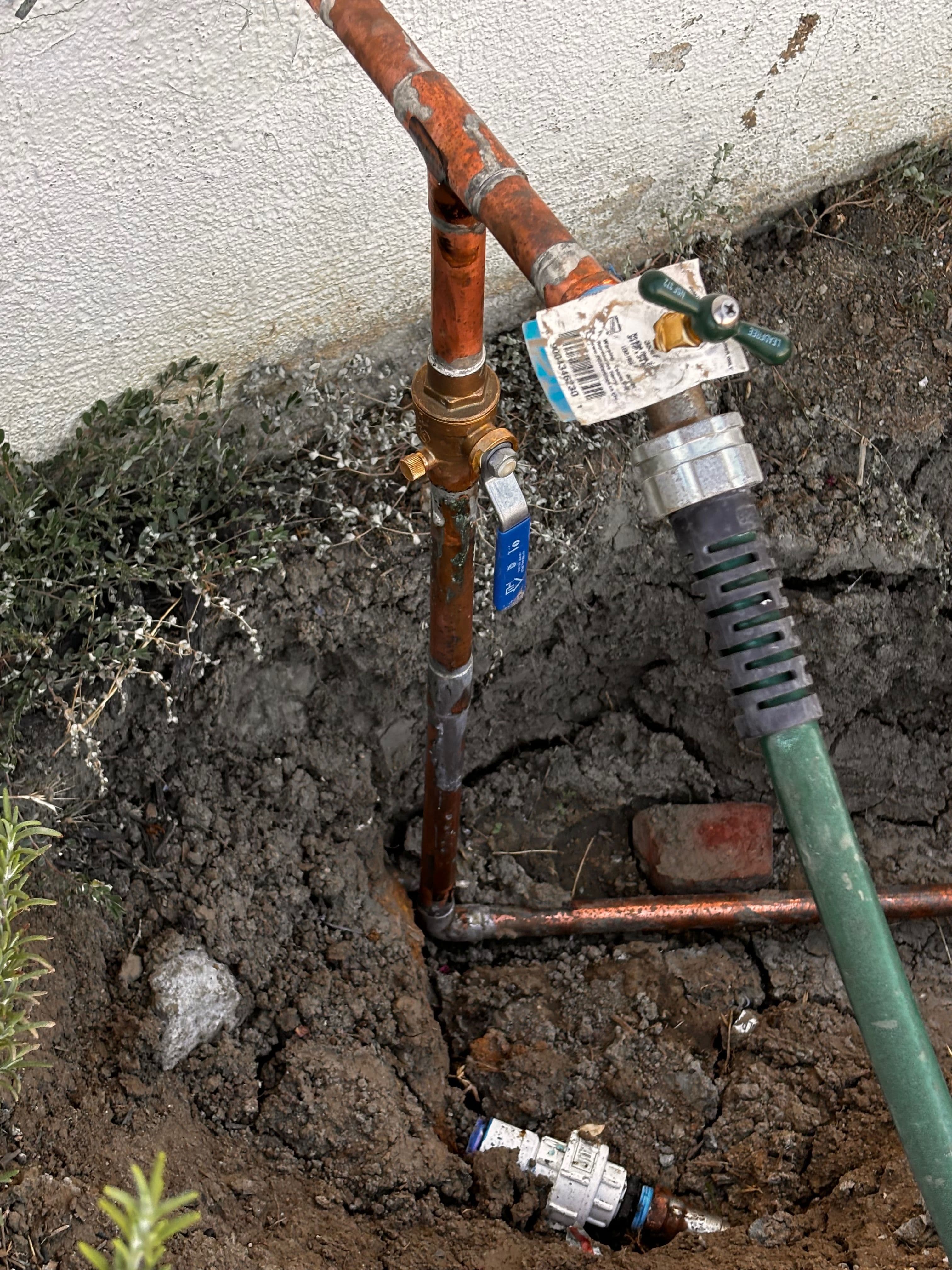 Copper water pipe with valves and a hose attachment in a dug-out garden area.