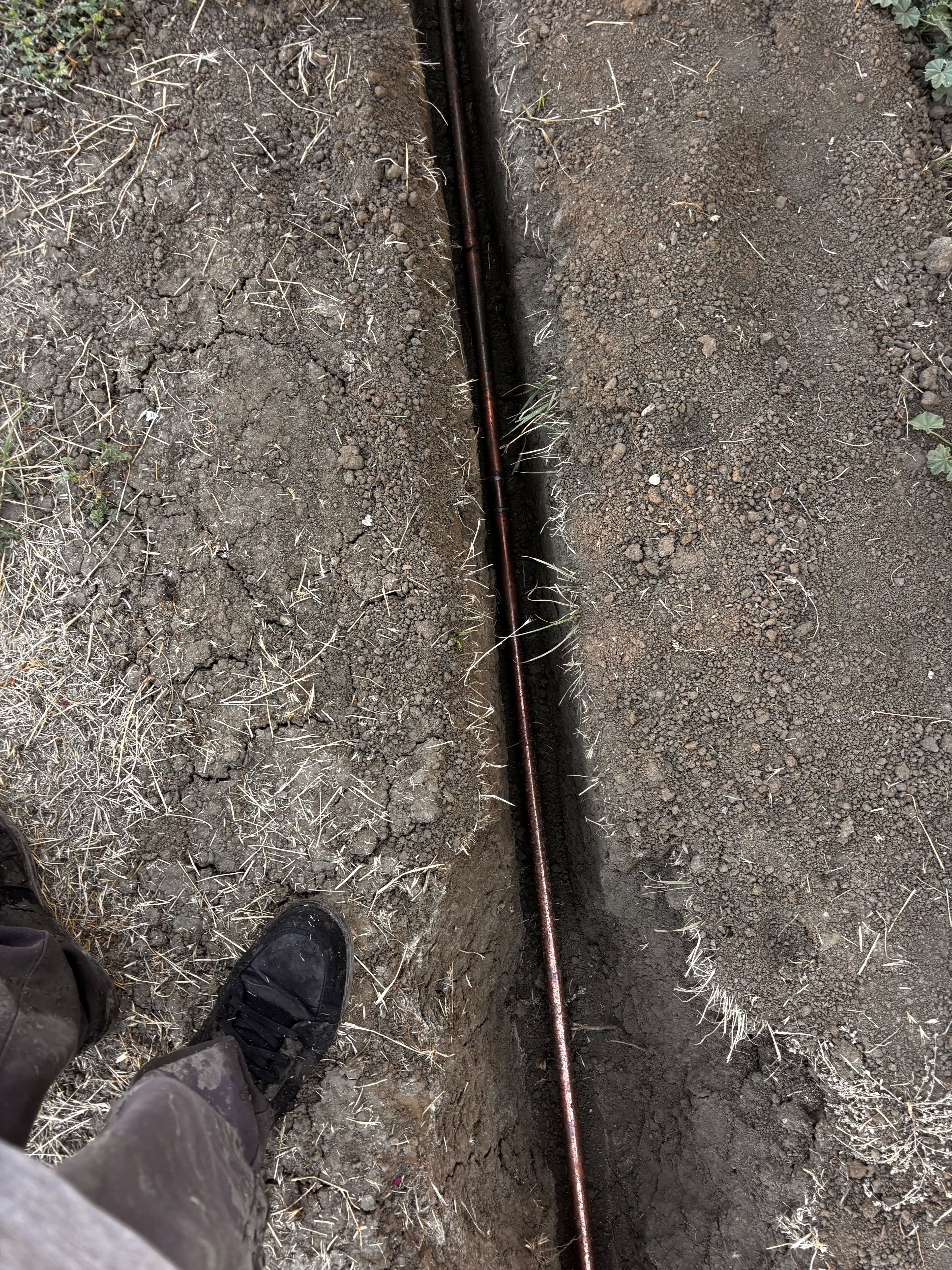 Trench revealing underground copper pipe, with a person's foot beside it on dry soil.