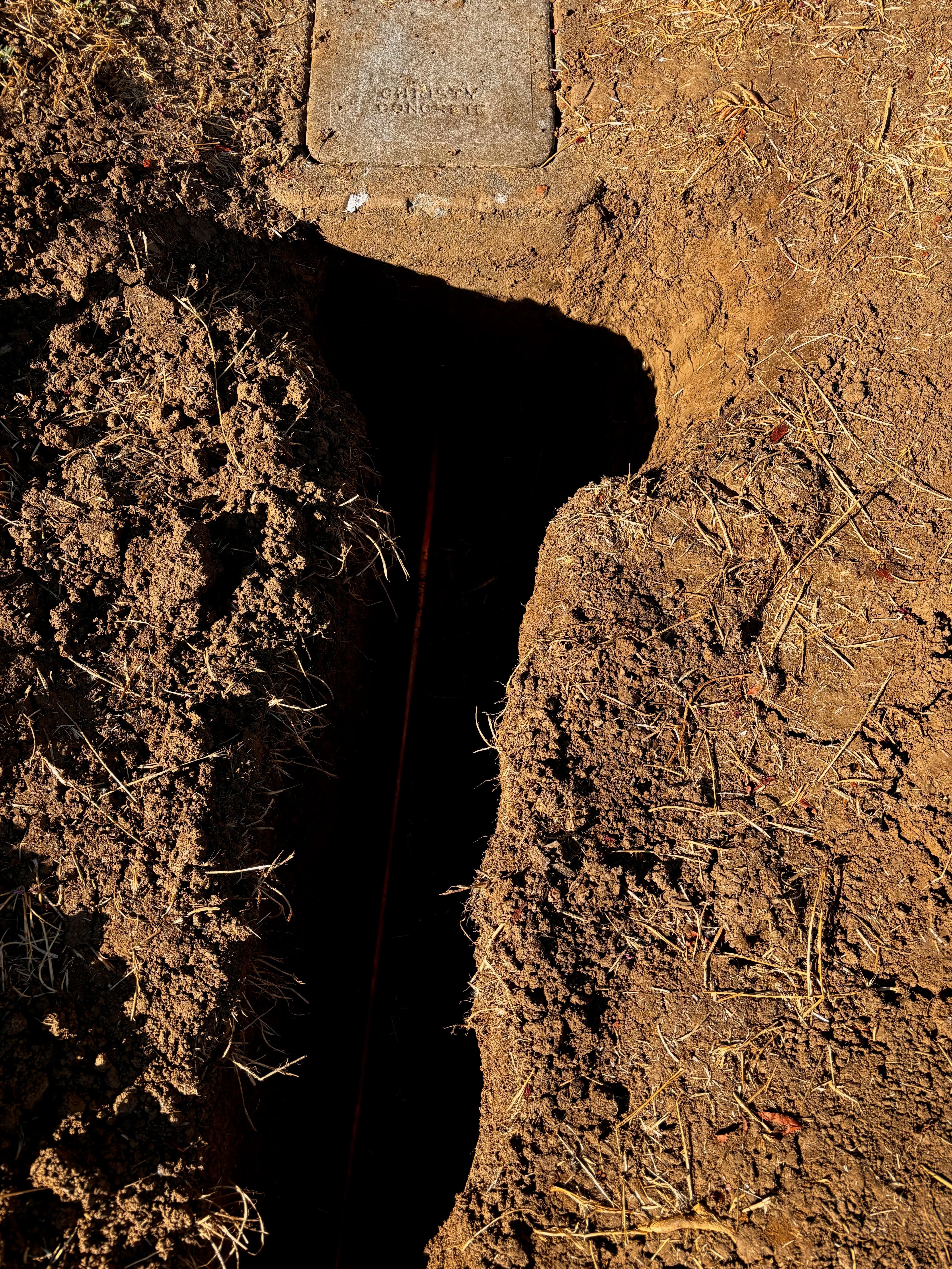 Exposed trench in soil revealing underground utilities, with a manhole cover nearby.