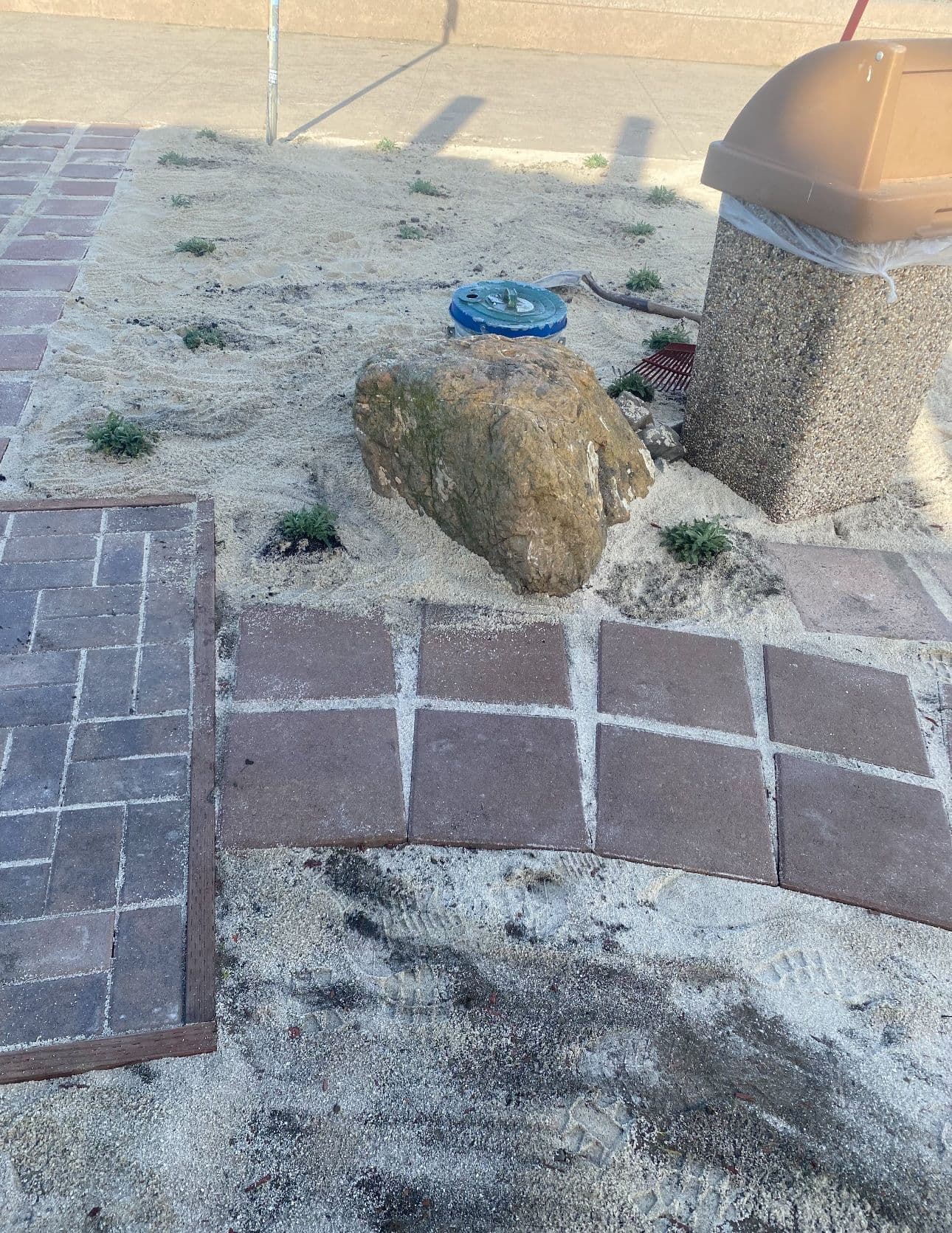 Pathway with brown pavers, sandy area, a rock, and a trash can in an outdoor setting.