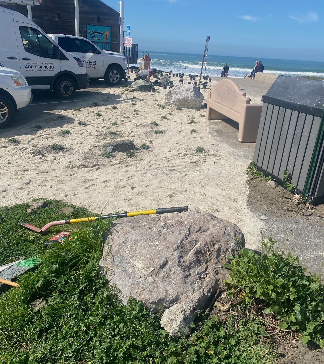 Beach scene with a rock, tools, and parked vehicles under a clear sky by the ocean.