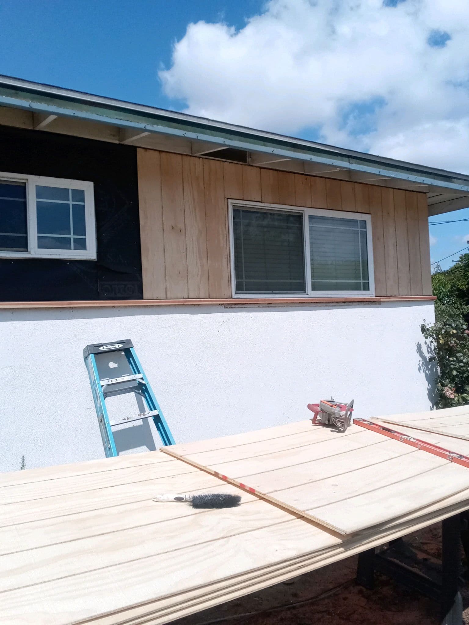 Siding installation on a house with plywood panels, ladder, and painting tools on a sunny day.