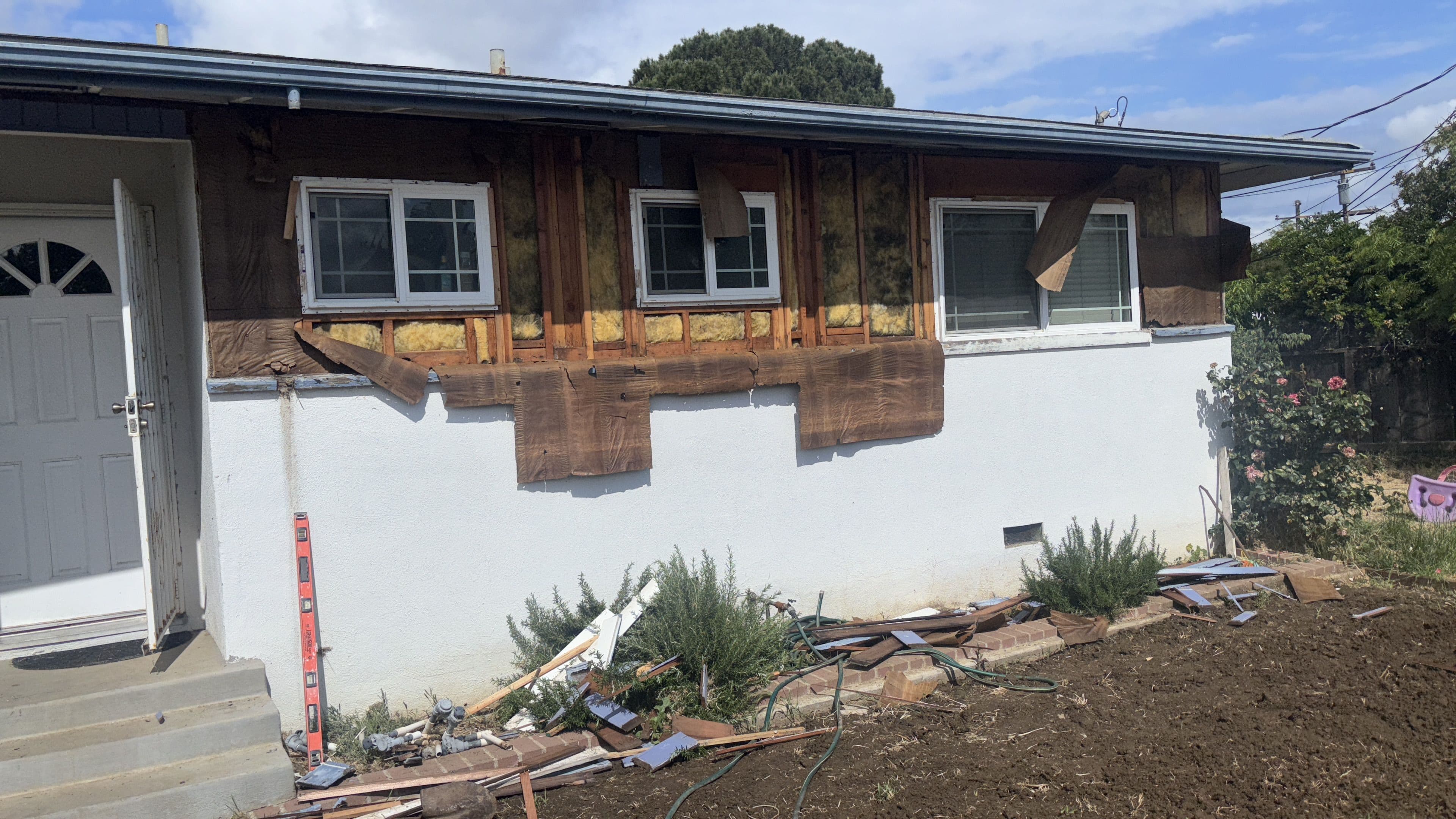 Home exterior in renovation with exposed wood, unfinished walls, and landscaping debris evident.
